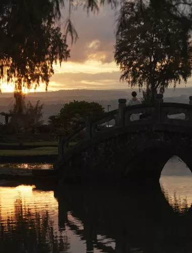 Liliuokalani Gardens A scenic view of Liliuokalani Gardens, a Japanese garden, at sunset with a traditional arched bridge over a calm pond.