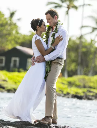 Honeymoons on the Island of Hawaii Bride and groom embracing on a rocky shoreline with greenery and palm trees in the background.