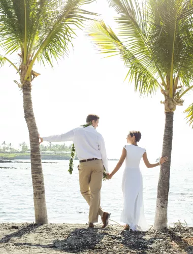 Weddings on the Island of Hawaii Couple holding hands under palm trees by the ocean, gazing into the distance on a sunny day.