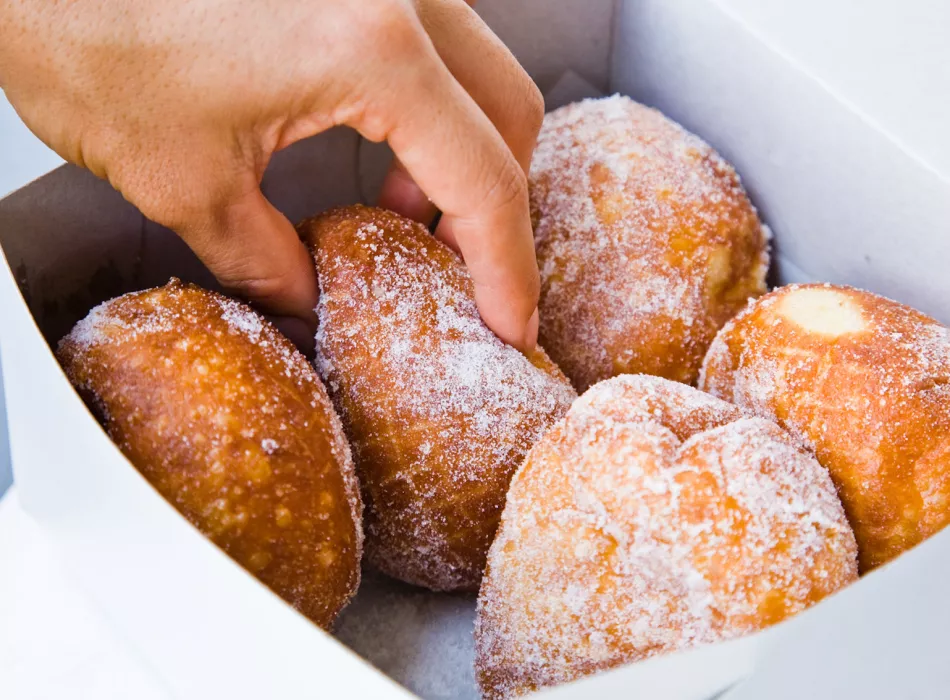 Close-up of a hand reaching for sugar-coated malasadas inside a box, showcasing the golden doughnuts.