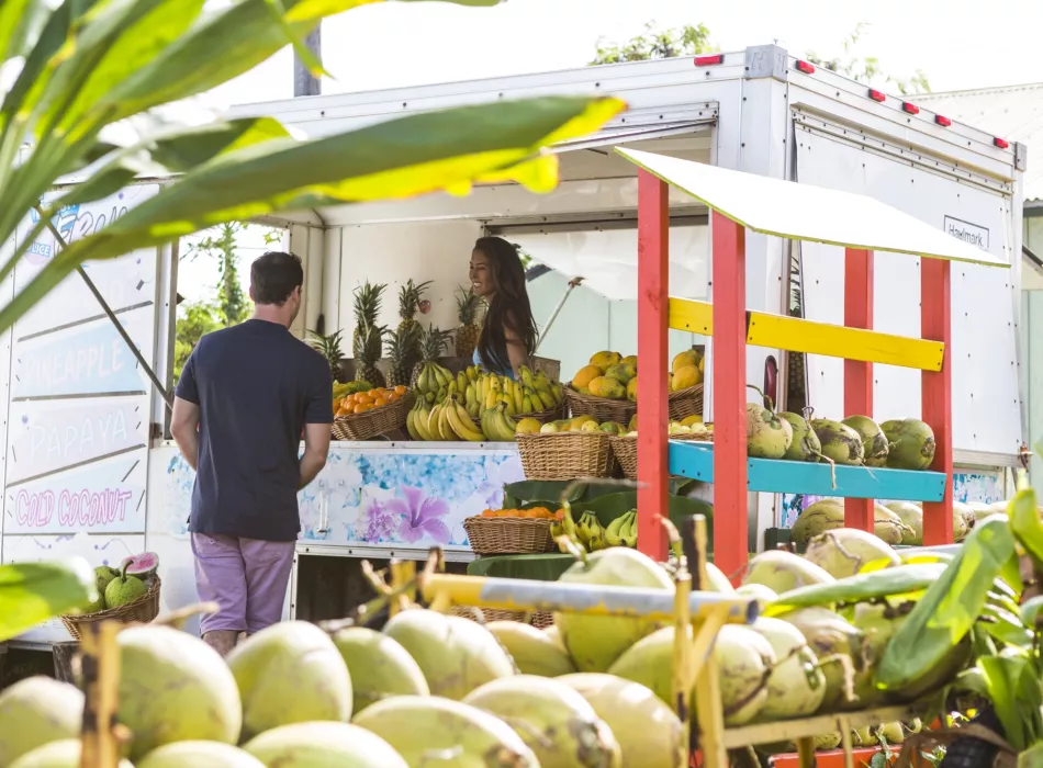 Fruit stand with baskets of bananas, pineapples, and coconuts, as a customer interacts with the vendor on a sunny day.