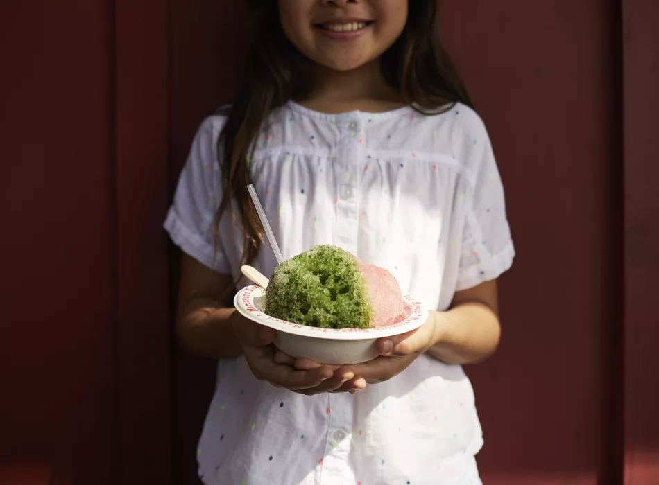 Smiling girl holding a bowl of colorful shave ice, topped with green and pink flavors, served with a spoon and straw.