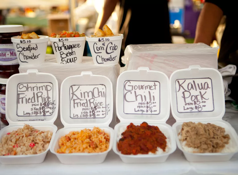 Display of Hawaiian plate lunches, including containers of kimchi fried rice, gourmet chili, and kalua pork, with soups in the background.