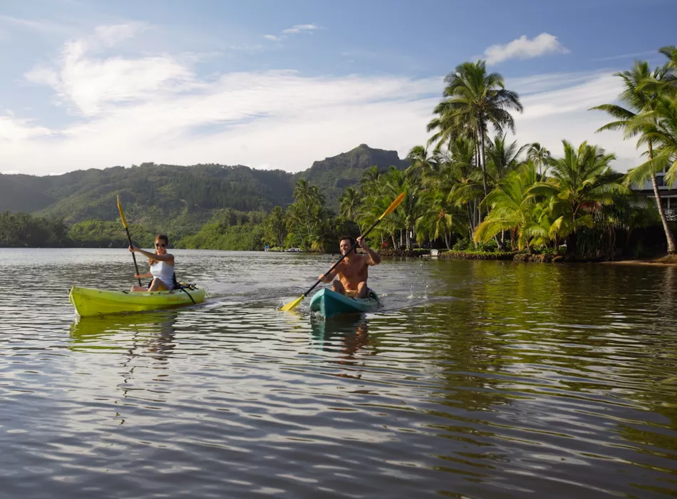 Two people kayaking on a calm river surrounded by tropical palm trees and lush greenery, with mountains in the background in Kaua‘i Parks.