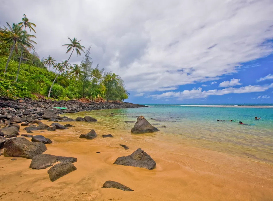 A pristine beach on Kaua‘i with golden sand, scattered rocks, turquoise water, and lush greenery under a partly cloudy sky.