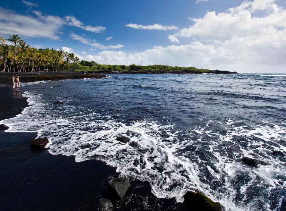A black-sand beach on the Island of Hawai‘i with waves meeting the shore, surrounded by lush greenery under a partly cloudy sky.