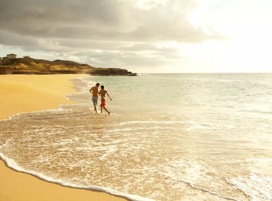 A wide sandy beach on Moloka‘i with gentle waves, two people walking along the shoreline, and a golden sky above.
