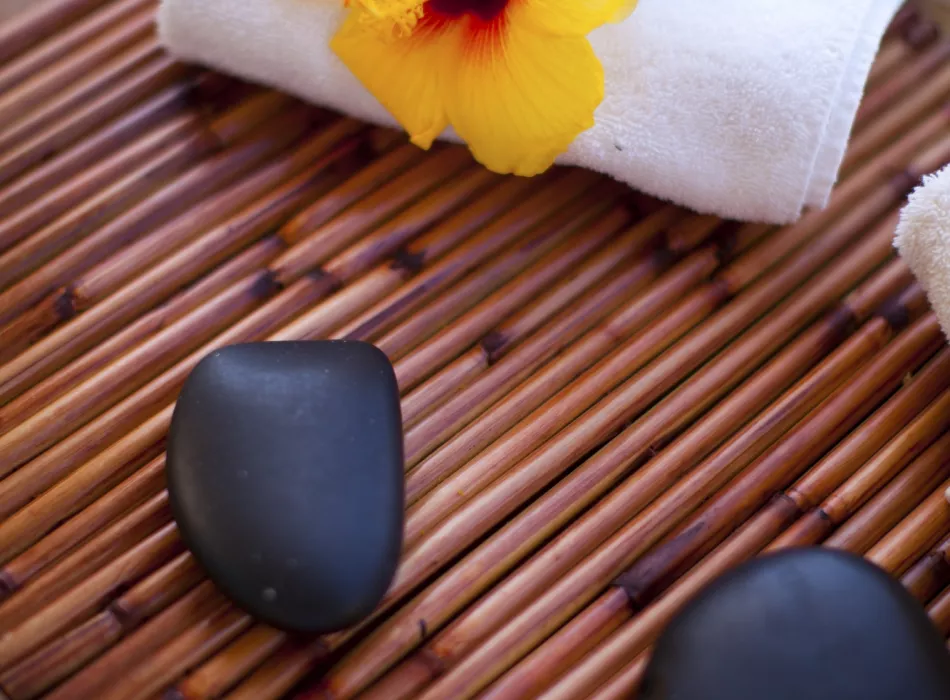Close-up of spa essentials on a bamboo mat, featuring black stones, a hibiscus flower, and soft white towels.