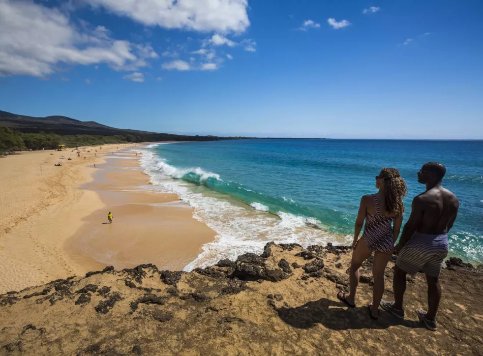 Two people stand on a rocky overlook enjoying the view of Mākena Beach, a long sandy shoreline with turquoise waves under a sunny sky.