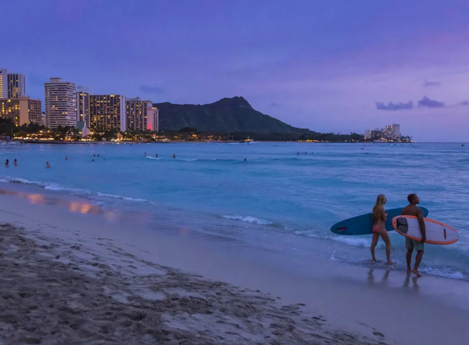 Two surfers with boards walking on the beach at sunset with Waikīkī cityscape and Diamond Head in the background.