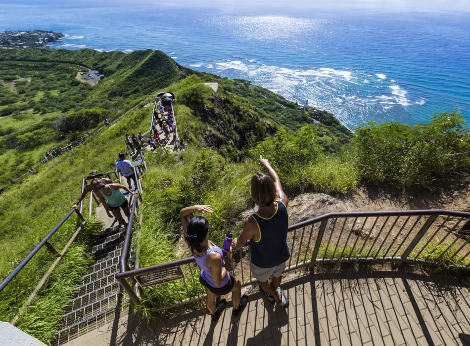 Visitors descending a scenic trail surrounded by lush greenery with expansive ocean views in the background.