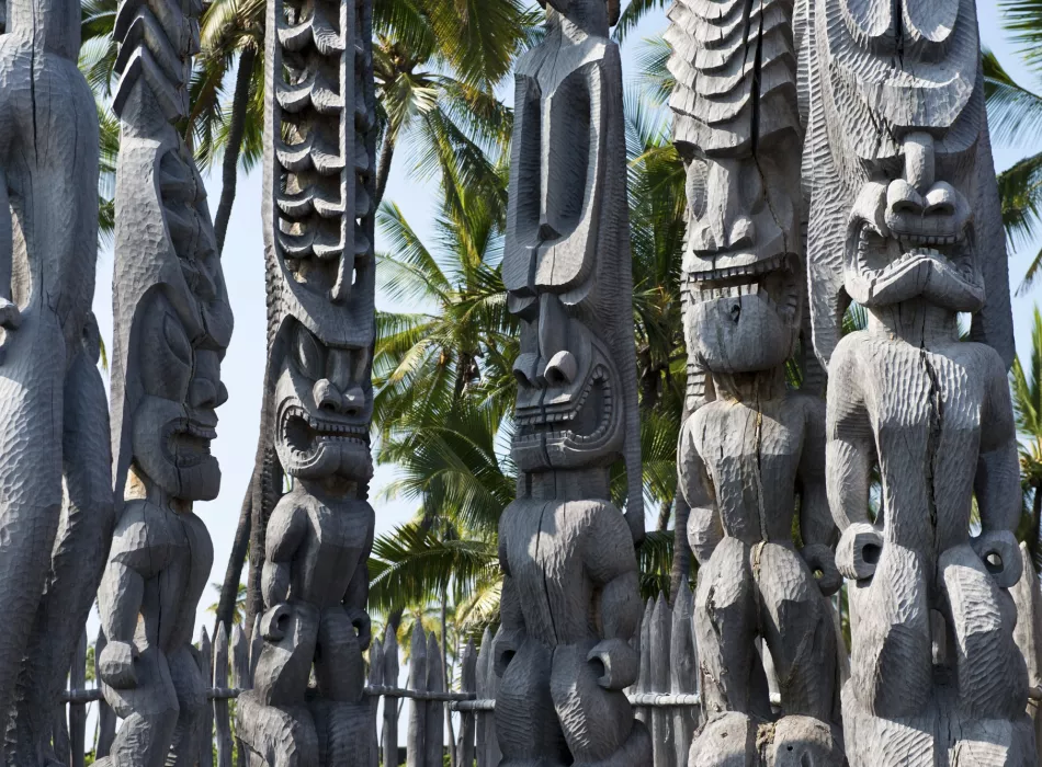 Traditional carved wooden statues at Pu‘uhonua o Hōnaunau National Historic Park, surrounded by palm trees under a clear sky.